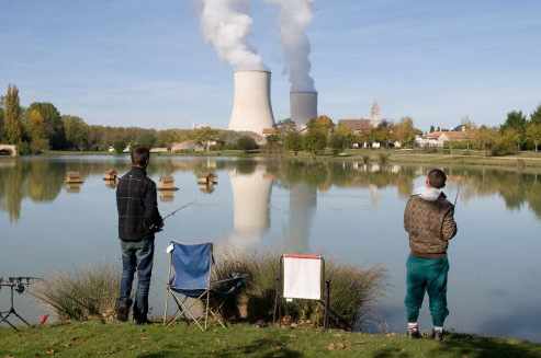 Dos personas pescando en un río al fondo una central nuclear