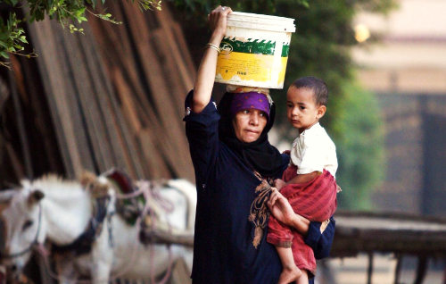 Una mujer con un niño en brazos y un cubo con agua en la cabeza