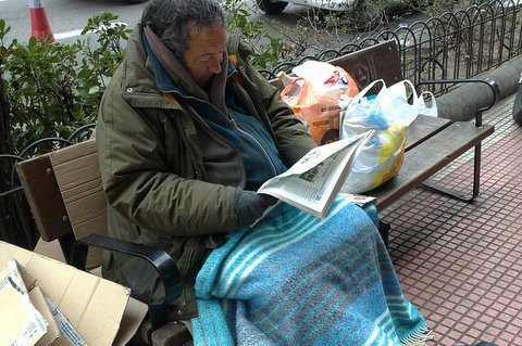 Un mendigo sentado en un banco leyendo el periódico
