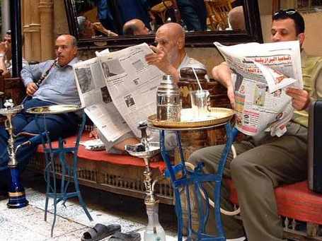 dos hombres leen sendos periódicos y un tercero fuma una pipa de agua