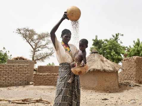 Una mujer africana con un bbebé en brazos echa semillas de un cuenco a otro