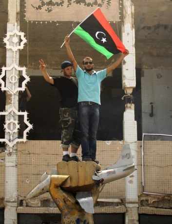 Dos libios con la bandera de los rebeldes encima de una escultura de gadafi