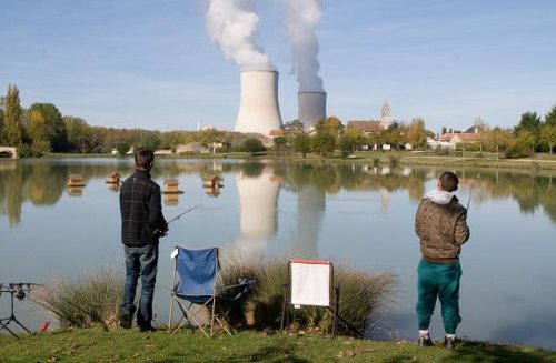 Dos hombres pescan en un río, al fondo una central nuclear