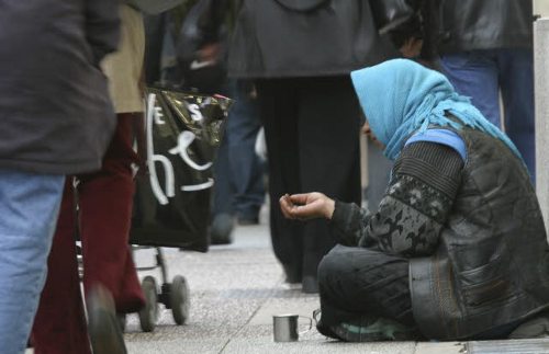 Mujer sentada en el suelo pidiendo entre personas que pasan