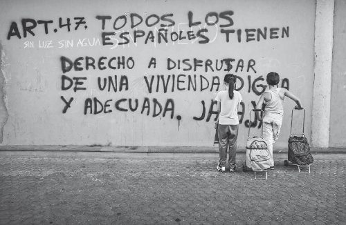 Unos niños mal vestido leen un grafitti en una pared que dice: Art. 47 Todos los españoles tienen derecho a disfrutar de una vivienda digna y adecuada. JA, JA, JA