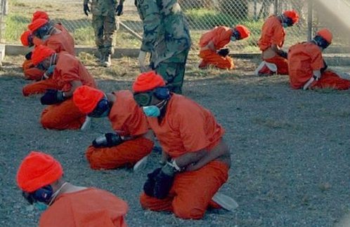 Los presos con mono naranja, gafas, máscaras de gas y guantes arrodillados en el suelo
