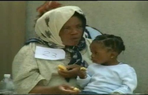 Mujer subsahariana con un número en el hombro con su niña en el regazo comen algún alimento
