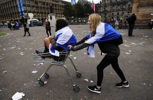 Una chica con una bandera de Escocia sobre los hombros empuja un carrito en el que va otra chica