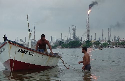 Dos hombres en una barca, al fondo una refinería